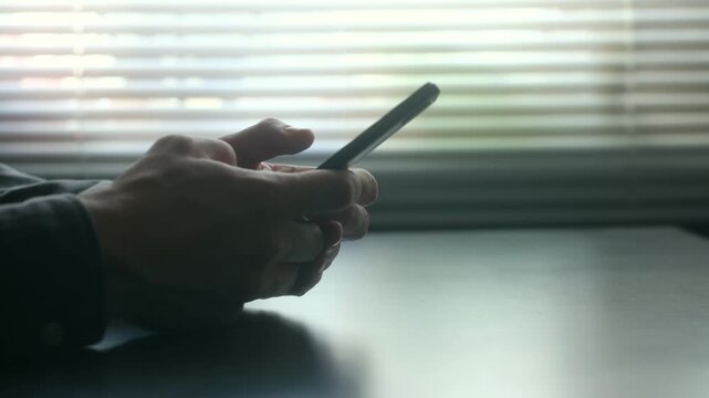 Male hands holding mobile phone typing message, texting, scrolling through social media while sitting at dark desk in front of window with blinds letting in daylight. Hands using smartphone at home