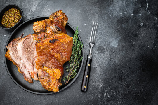 Crispy Schweinshaxe showing blistered skin, rendered fatcap and tender sliced interior, plated on dark textured background with herbs.