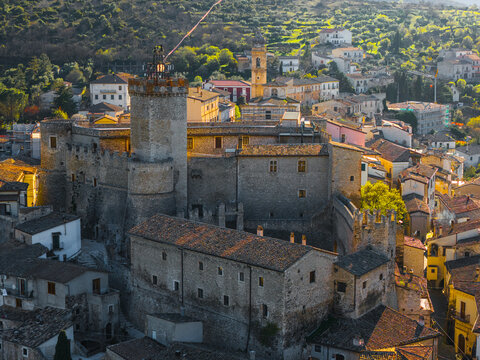 Aerial view of the medieval Castello Piccolomini with its tall stone tower and surrounding historic rooftops under warm evening light, Capestrano, Abruzzo, Italy.