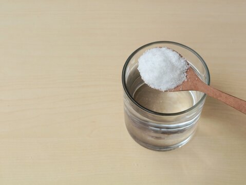 Glass of salt water solution with salt on wooden spoon on wooden table. Concept of saline solution, home remedies, healthy minerals, or cooking ingredients