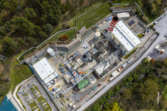 Aerial view of an industrial chemical plant with a tall red and white striped chimney, complex piping systems, and storage tanks nestled among green hills in Bussi sul Tirino, Abruzzo, Italy.