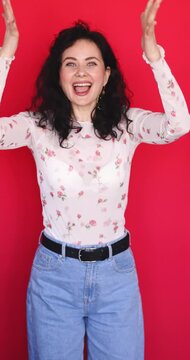 Happy young woman raising hands in celebration or surprise, laughing on red background. Joyful brunette girl in floral blouse expressing success, happiness and positive emotions indoor