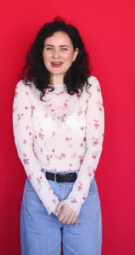 Coy and charming young woman with curly hair looking at camera and smiling modestly on red background. Sweet brunette girl in floral sheer blouse posing with shy and friendly facial expression indoor