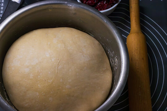 A bowl with dough on a kitchen countertop, a dough roller and a bowl with sour cherry is nearby. 