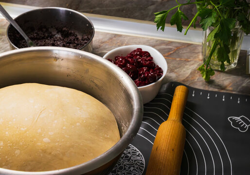A bowl with dough on a kitchen countertop, a dough roller and a bowl with sour cherry is nearby. 