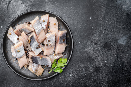 Pickled herring chunks cured in salt and brine served with spices and herbs. Traditional seafood on dark textured background.