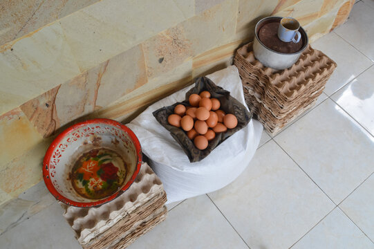 Looking down from a quarter-left angle at a bowl of fresh eggs, poultry feed sacks, and stacked plastic egg crates arranged on a tiled residential entryway.