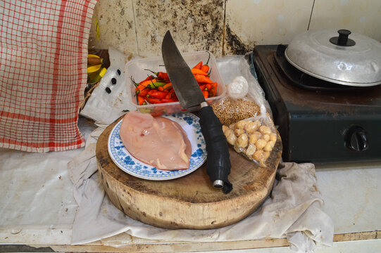 A high angle shot showing raw chicken pieces, red chili, garlic, and traditional spices next to a large machete knife on a wooden board in a domestic kitchen.
