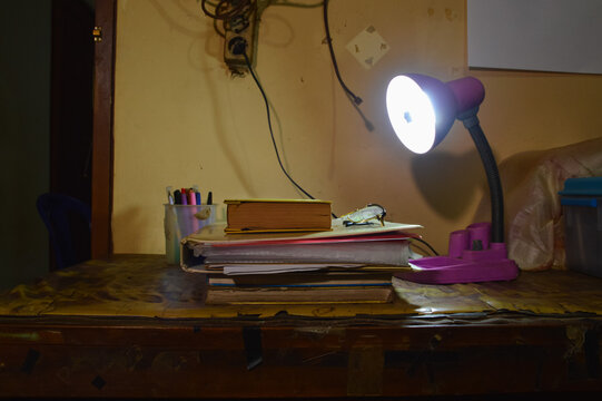 A straight eye level shot of a study desk featuring a neat stack of textbooks, reading glasses, a modern desk lamp, and a stationery organizer in a home office setting.