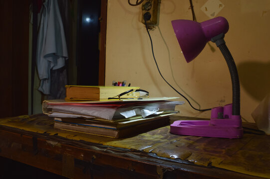 A quarter right perspective of a residential workspace showing a pile of books, black reading glasses, and an adjustable desk lamp on a wooden table surface.