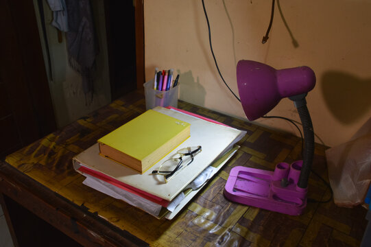 A quarter right high angle shot looking down at a stack of academic books, eyeglasses, and office supplies arranged neatly on a desk under indoor lighting.