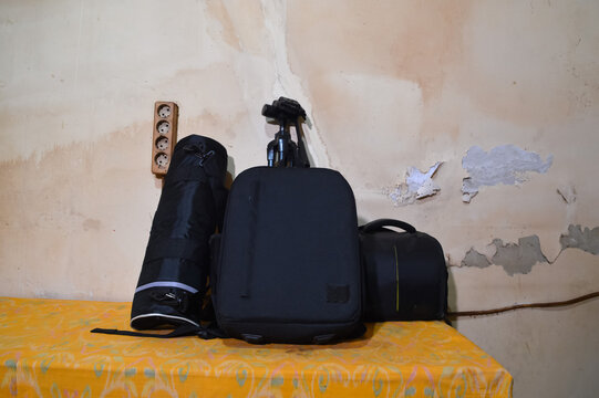 A straight eye level view of various photography bags, including a backpack and a shoulder bag, leaning against a wall on a yellow covered table inside a room.