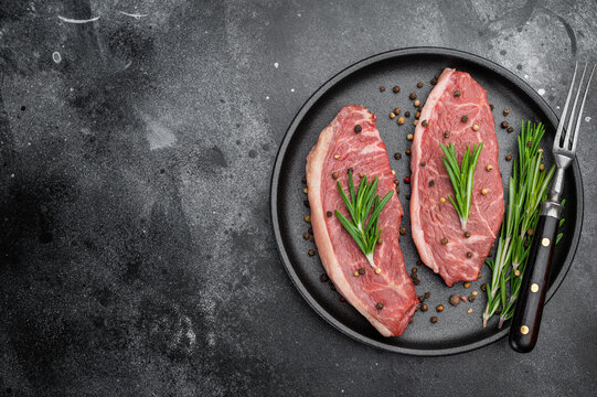 Raw picanha beef steaks also known as top sirloin cap and rump cap with rosemary and pepper on black plate, dark stone background