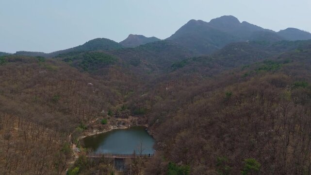 Cinematic aerial view of a calm mountain lake surrounded by leafless trees in early spring. Muted tones and still water create a serene, moody landscape capturing seasonal transition.
