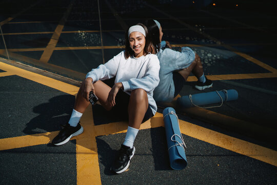 Woman poses casually while holding smartphone, seated in sunlight against reflective wall, yoga mat unrolled nearby, embodying balance between mobile presence and body awareness.