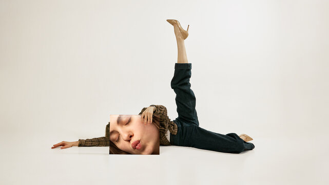 Young woman lying with leg raised holding printed face panel minimal fashion studio portrait. Concept of bold posing, high fashion attitude, body styling and contemporary editorial aesthetics.