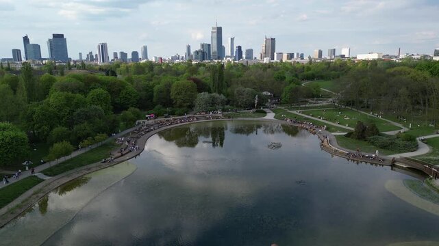 Aerial view of urban park with lush greenery and a serene pond reflecting city skyline, showcasing people enjoying leisure activities in a vibrant natural setting