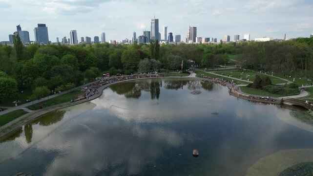 Aerial view of a vibrant urban park with a serene pond surrounded by lush greenery and city skyline, showcasing people enjoying leisure activities in the background