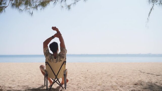 Asian man relaxing on beach chair with arms raised facing the sea, enjoying freedom lifestyle during summer vacation, solo travel social detox slow life moment by ocean, outdoor leisure with blue sky