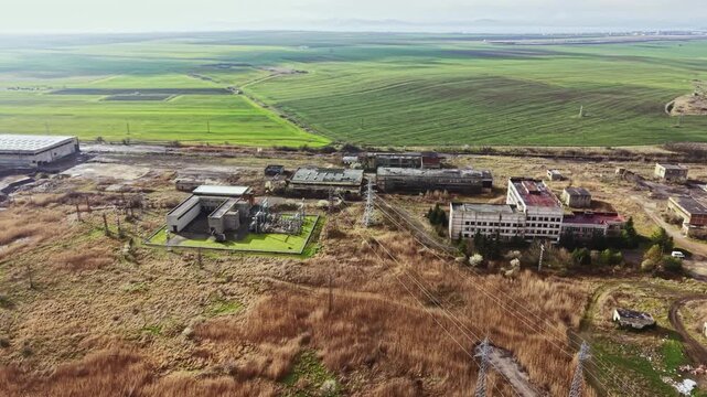 Aerial view shows abandoned buildings and overgrown fields in a rural area. Power lines stretch across the landscape under a clear sky. The scene captures emptiness and nature reclaiming space.