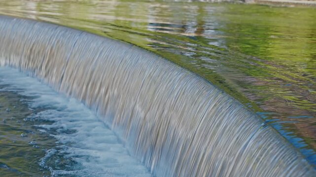 A serene close-up shot of water flowing in a smooth, glass-like curtain over a curved stone edge at Heisei Teien Garden. Bubbles and foam form at the base in a tranquil setting.