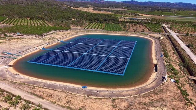Floating solar panel array installed on agricultural irrigation reservoir surrounded by orchards and farmland, blue photovoltaic modules on pontoon structure reducing water evaporation