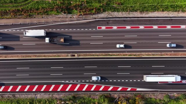 Top-down view of a dual carriageway highway featuring white dashed lane markings, red and white chevron road markings, with cars and trucks in motion