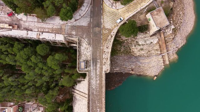 Overhead view of Benageber dam on the Turia river in Valencia, Spain, revealing turquoise reservoir water, concrete dam wall, rocky cliffs and surrounding pine forest