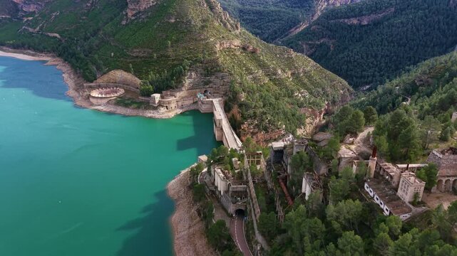 Benageber dam on the Turia river in Valencia, Spain, with turquoise reservoir waters, ruined industrial buildings, and forested mountain canyon landscape