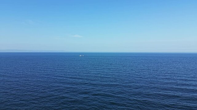 A broad view of the ocean shows smooth water under a cloudless sky. The scene captures a boat in the distance, making its way across the water in bright daylight.