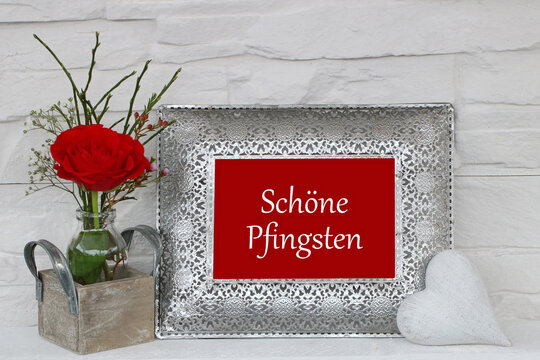 Elegante Pfingstgr&uuml;&szlig;e: Vase mit roter Rose und dem Schriftzug Sch&ouml;ne Pfingsten auf einem Bilderrahmen.
