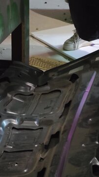 A person in a black jacket, red gloves, and white shoes stands on a disassembled car hood, pounding a keyboard on the floor below with a wooden baseball bat in a garage setting.