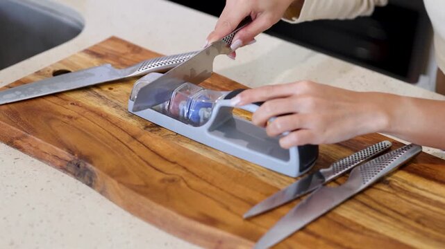 Close up of hands sharpening a kitchen knife on a multi stage sharpener on wooden cutting board.
