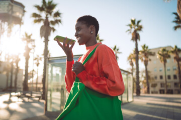 Wide shot of Black woman seated on a sandy beach holding green smartphone and smiling, showing beachside mobile use, digital browsing, and relaxed tech interaction with media. © BullRun