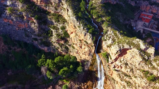 Water from a waterfall cascades down rocky cliffs surrounded by trees. A building is visible in the background. The scene is captured in daylight, showing the natural beauty of the area.