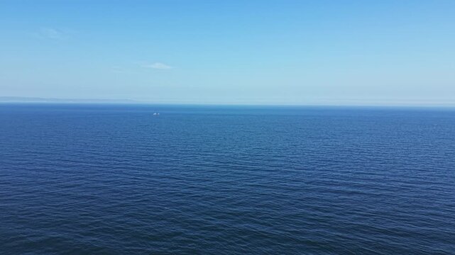 A large expanse of ocean stretches beneath a clear blue sky during daytime. The water has gentle waves and smooth surface. A small boat is visible in the distance.