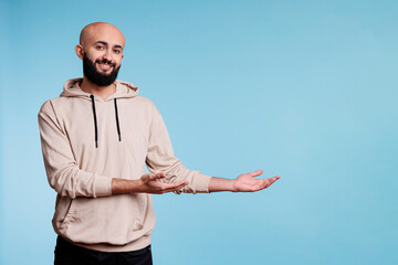 Naklejka premium Cheerful smiling arab man pointing to left while looking at camera. Happy joyful young person advertising product and showing to side portrait while posing on blue background