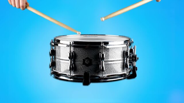 A close-up shot of a metallic snare drum being struck by drumsticks against a vibrant blue studio background. This rhythmic footage is perfect for music production, band advertisements, and creative p