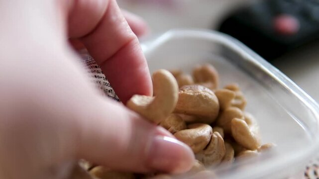 Close-up of cashew nuts in a plastic container. A hand picks up the nuts, showing texture and freshness. Simple healthy snack moment with natural lighting and minimal composition.