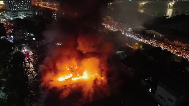 Wide aerial cityscape showing major fire in Hanoi near river, flames and smoke dominating skyline while surrounding buildings remain lit.