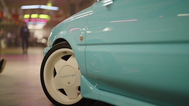 Close-up shot of a classic custom car with unique white wheels spinning in a dimly lit underground parking lot, with neon lights and people walking in the blurry background