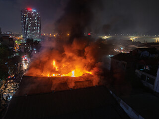 Close aerial shot of rooftop engulfed in flames in Hanoi, heavy smoke rising while nearby high-rise building and riverfront remain visible at night. © Hanoi Photography
