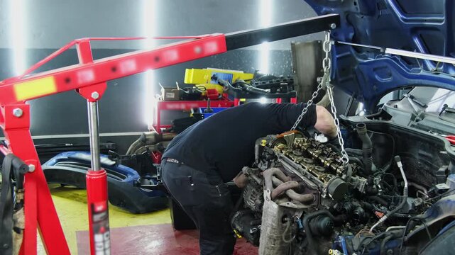 Skilled mechanic in workwear dismantles car engine with hoist at service station slow motion. Worker disconnects vehicle motor in repair shop