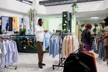 Fototapeta premium Clothes shop african american employee hanging shirt on rack. Fashion garment showroom assistant checking formal wear while working in store and choosing apparel for customer