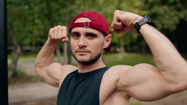 A fit man shows off his muscles in a park. He flexes his biceps and looks at the camera with a serious expression. This video evokes feelings of strength, health, and dedication to fitness.