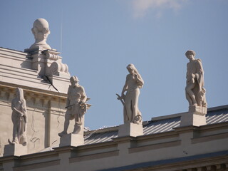 Obraz premium Row of classical statues standing on the rooftop of a historic building, silhouetted against a bright blue sky with soft clouds. Taken in front of the Hungarian Parliament in Budapest.