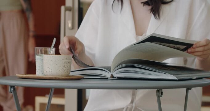 A young woman in a white robe engaged in reading a large, open book at a table. A coffee cup and saucer sit beside her, depicting a relaxed, quiet morning.
