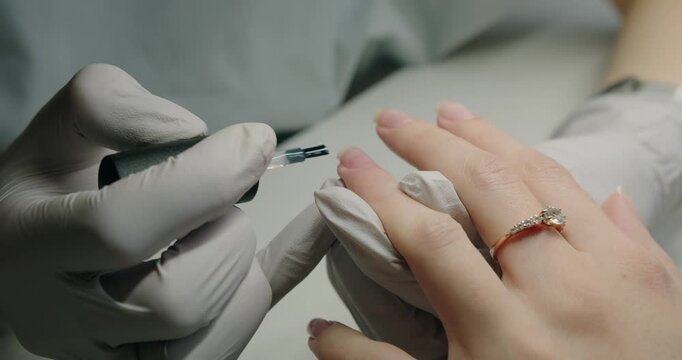 Woman salon visitor waits for refined and eye-catching result after nail procedure. Manicure master hands in gloves applying gel polish on client nail at beauty salon. Professional care female hands.