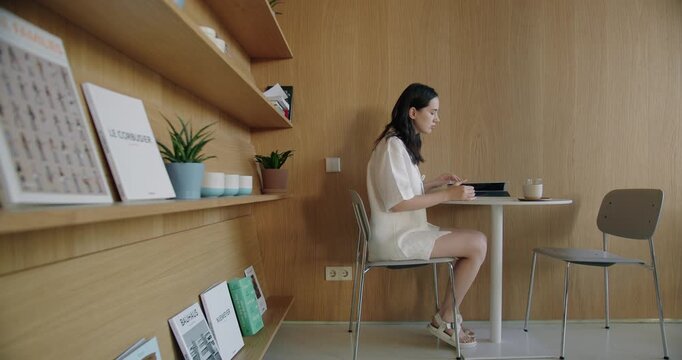 A focused young woman works on a tablet in a serene setting, surrounded by books and plants in a modern wooden interior. concentration and calm in a professional environment.