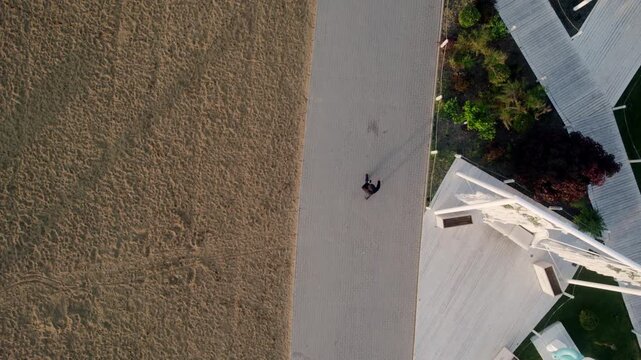Top-down aerial view of a solitary person walking along a modern promenade that separates a sandy beach from a green park area with contemporary white architectural elements and wooden decks